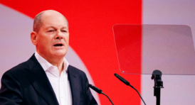 Bundeskanzler Olaf Scholz bei einer Rede auf einem Podium vor einem roten Hintergrund im Querformat.